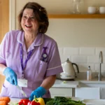 A smiling woman in a light purple nurse uniform and blue gloves prepares vegetables in a kitchen. Carrots, peppers, and greens are on the worktop. Shelves and a kettle are visible in the background.