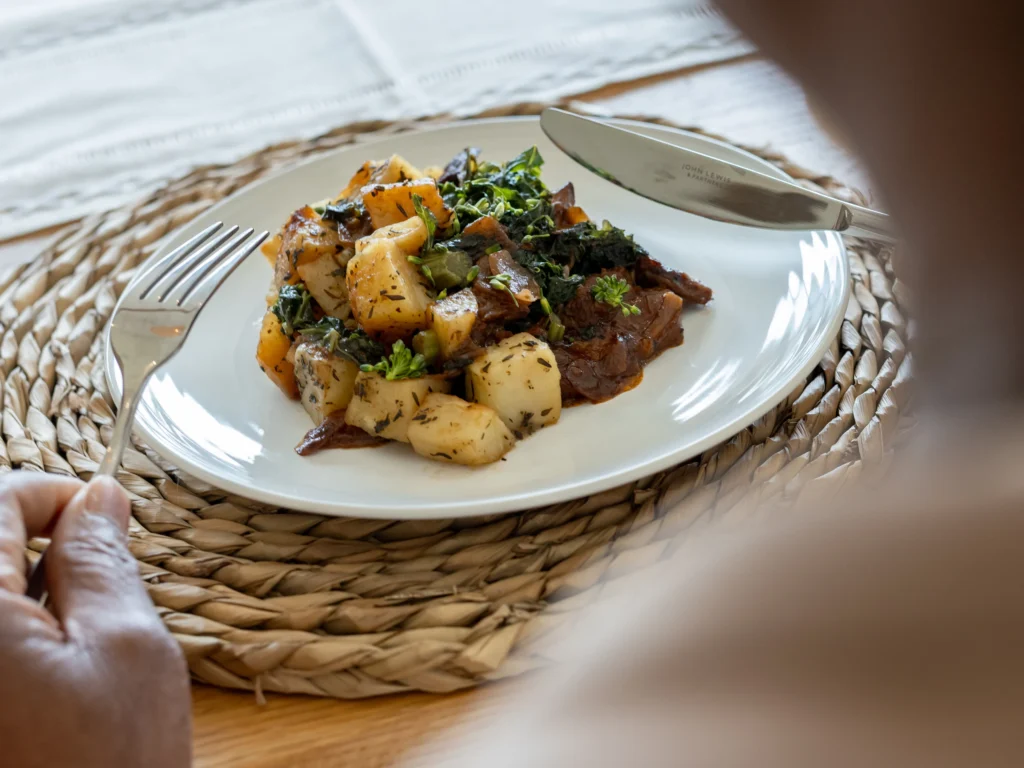 A person holding a fork and knife is about to eat a plate of roast potatoes, leafy greens, and pieces of meat, served on a white plate atop a woven placemat.