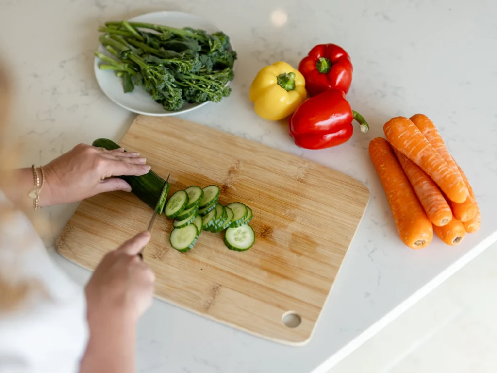 A person slices a cucumber on a wooden chopping board. Nearby on a white worktop are carrots, red and yellow peppers, broccolini, and a white plate.