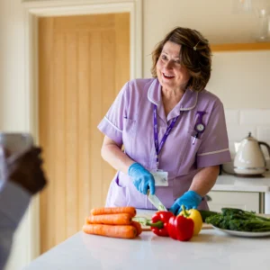 A smiling female carer in a purple uniform and blue gloves slices vegetables, including carrots and peppers, on a kitchen worktop, while talking to someone holding a mug.