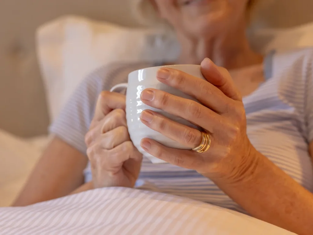 A person with light skin and gold rings holds a white mug whilst relaxing in bed, wearing a striped shirt. The scene appears cosy and peaceful. The person's face is partially out of focus.