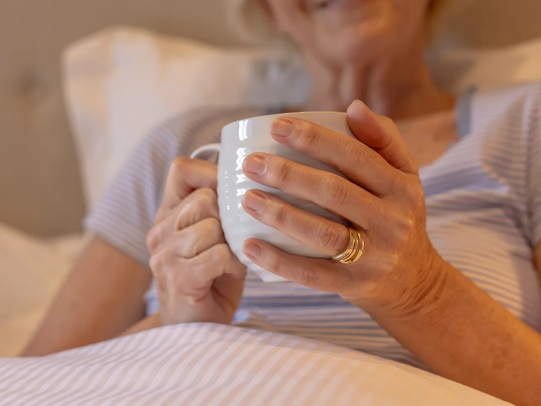 A person with light skin and gold rings holds a white mug whilst relaxing in bed, wearing a striped shirt. The scene appears cosy and peaceful. The person's face is partially out of focus.