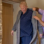 A younger man helps an older man put on a grey dressing gown in a cosy room with stone and wooden walls. The older man wears navy pyjamas and looks down as he is being assisted.