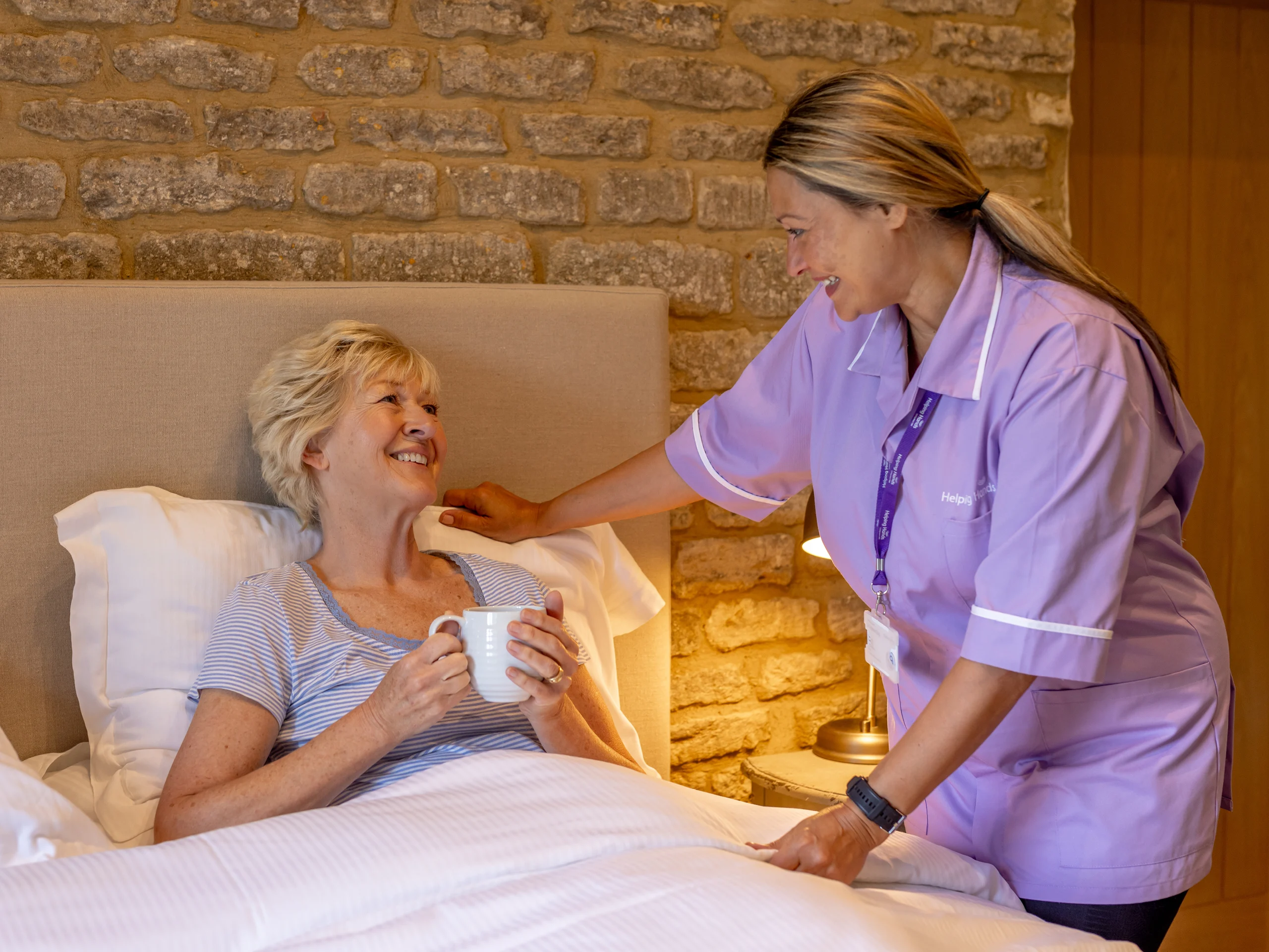 A carer in a purple uniform smiles and gently touches the shoulder of an elderly woman sitting up in bed, holding a mug, in a cosy room with stone walls.