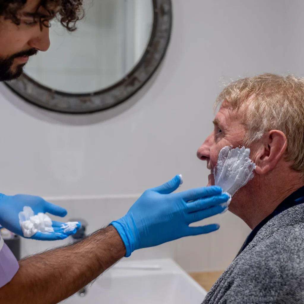 A person wearing blue gloves applies shaving foam to the face of an older man sitting in a bathroom, with a round mirror hanging on the wall in the background.