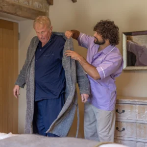 A carer helps an older man put on a grey dressing gown in a cosy bedroom. The carer wears a light purple uniform, and the man is dressed in navy blue pyjamas. A wooden chest of drawers and a mirror are visible in the background.