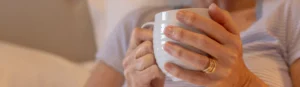 A close-up of a person wearing a striped shirt holding a white mug with both hands, showcasing gold rings on their fingers. The background is softly blurred, suggesting a cosy indoor setting.