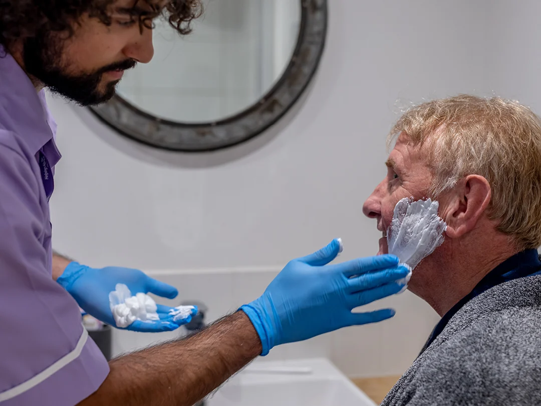 A carer in a purple uniform and blue gloves applies shaving foam to the face of an elderly man sitting by a washbasin, preparing him for a shave. A round mirror hangs on the wall behind them.