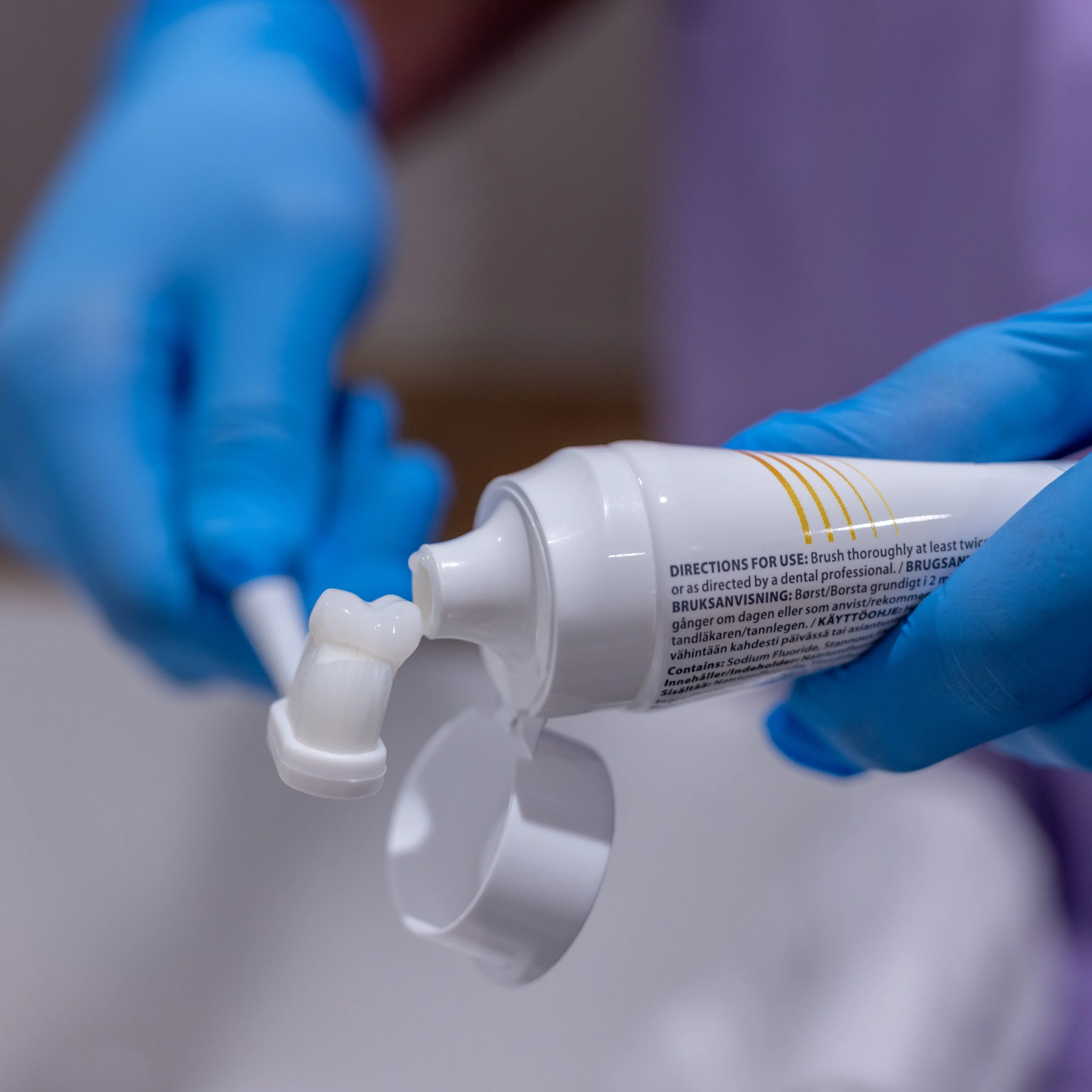 A person wearing blue gloves squeezes toothpaste onto a toothbrush from a white tube, with the cap open and visible instructions on the tube.