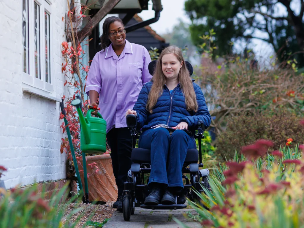 A smiling woman in a wheelchair is outside in a garden, accompanied by a carer holding a green watering can. Both appear happy, enjoying the garden path lined with plants and flowers.