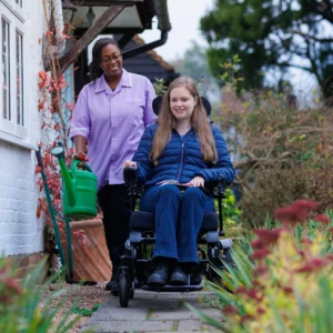 A smiling woman in a wheelchair moves along a garden path, while another woman in a purple shirt walks behind her holding a green watering can. The background shows plants and shrubs near a white building.