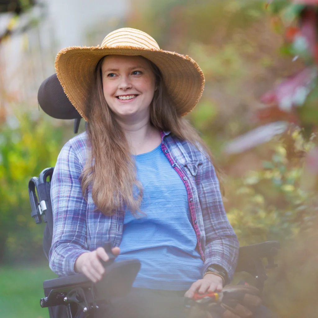 A young woman with long hair, wearing a straw hat, blue shirt, and checked shirt, smiles while sitting in a motorised wheelchair outdoors among greenery.