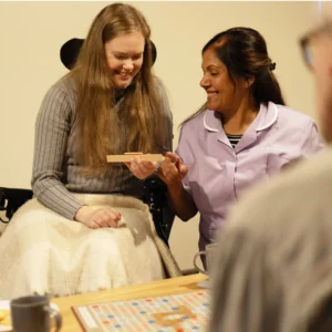 A woman in a wheelchair smiles while holding Scrabble tiles, sitting next to a carer in a purple uniform who is also smiling. A Scrabble board and mugs are on the table in front of them.