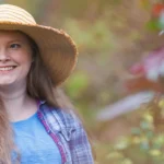 A woman with long hair wearing a wide-brimmed hat, blue shirt, and checked overshirt smiles whilst sitting in a wheelchair outdoors in a garden setting.