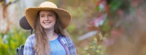 A woman with long hair wearing a wide-brimmed hat, blue shirt, and checked overshirt smiles whilst sitting in a wheelchair outdoors in a garden setting.