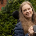 A woman with long light brown hair sits outdoors in a wheelchair, smiling and holding a grey mug. A blurred person is in the foreground, and greenery is in the background.