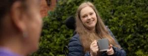 A woman with long light brown hair sits outdoors in a wheelchair, smiling and holding a grey mug. A blurred person is in the foreground, and greenery is in the background.