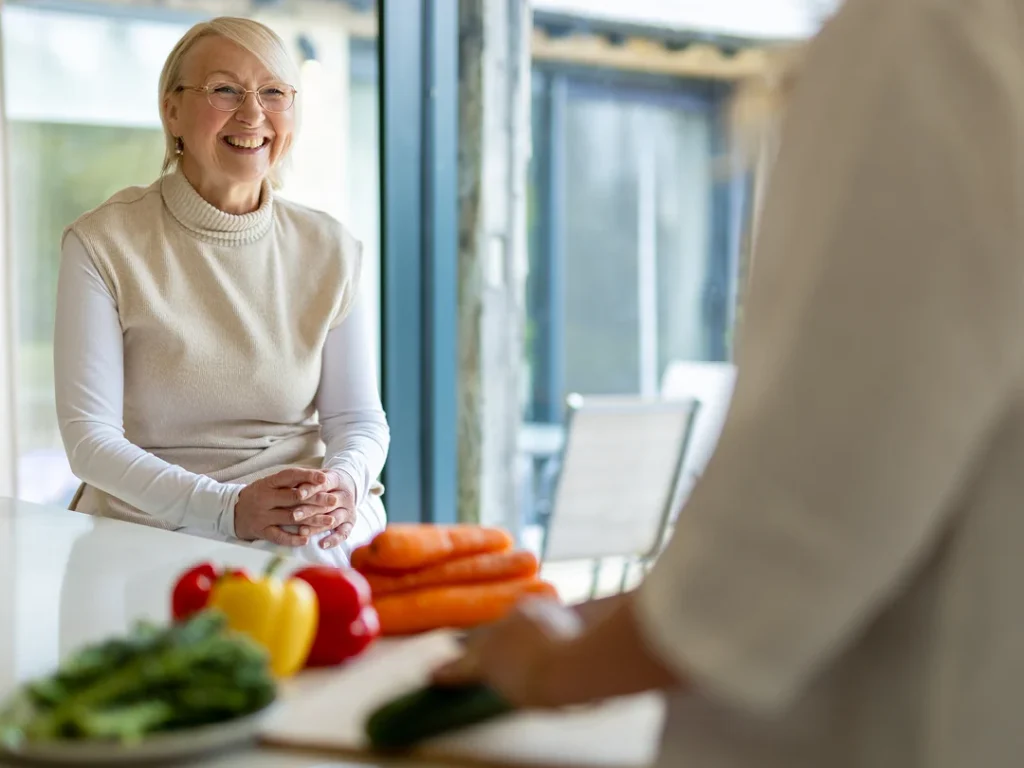 An older woman with glasses smiles whilst sitting at a kitchen worktop. Fresh vegetables, including carrots, peppers, and greens, are on the worktop, and another person is slicing a cucumber in the foreground.