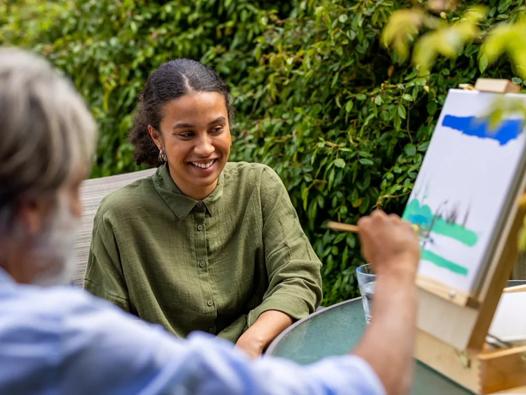 A woman in a green shirt smiles whilst sitting at an outdoor table, watching as another person paints a landscape on a canvas, surrounded by lush greenery.