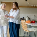 Two women smile at each other as they walk arm-in-arm in a bright kitchen. One uses a walking stick. Vegetables and flowers are on the worktop, creating a warm, cheerful atmosphere.