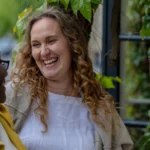 Two people stand outside by a wall covered in green leaves, smiling warmly at each other. One has curly hair and wears a light top, the other wears glasses and a yellow jacket. The atmosphere is cheerful and friendly.