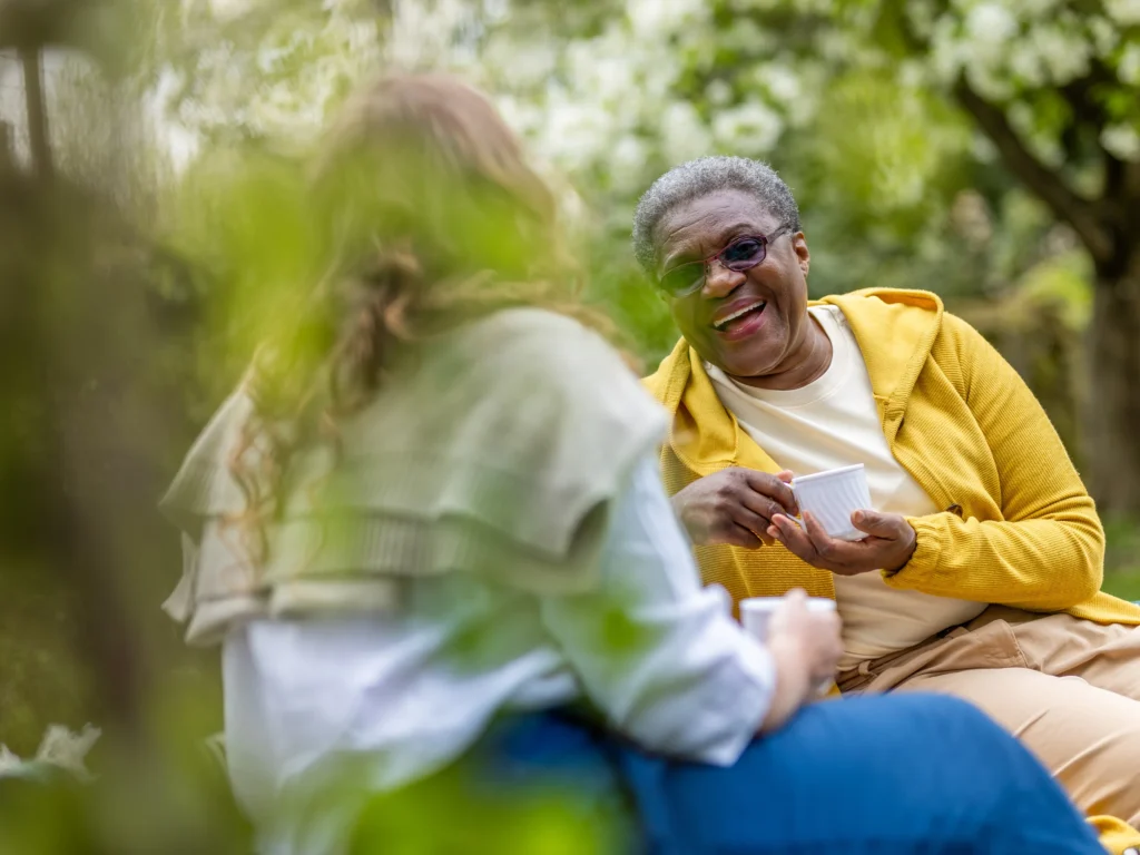 Two women sit outdoors, chatting and smiling while holding mugs. One woman wears a yellow hoodie and glasses, laughing warmly, while the other faces away, partially blurred by green foliage in the foreground.