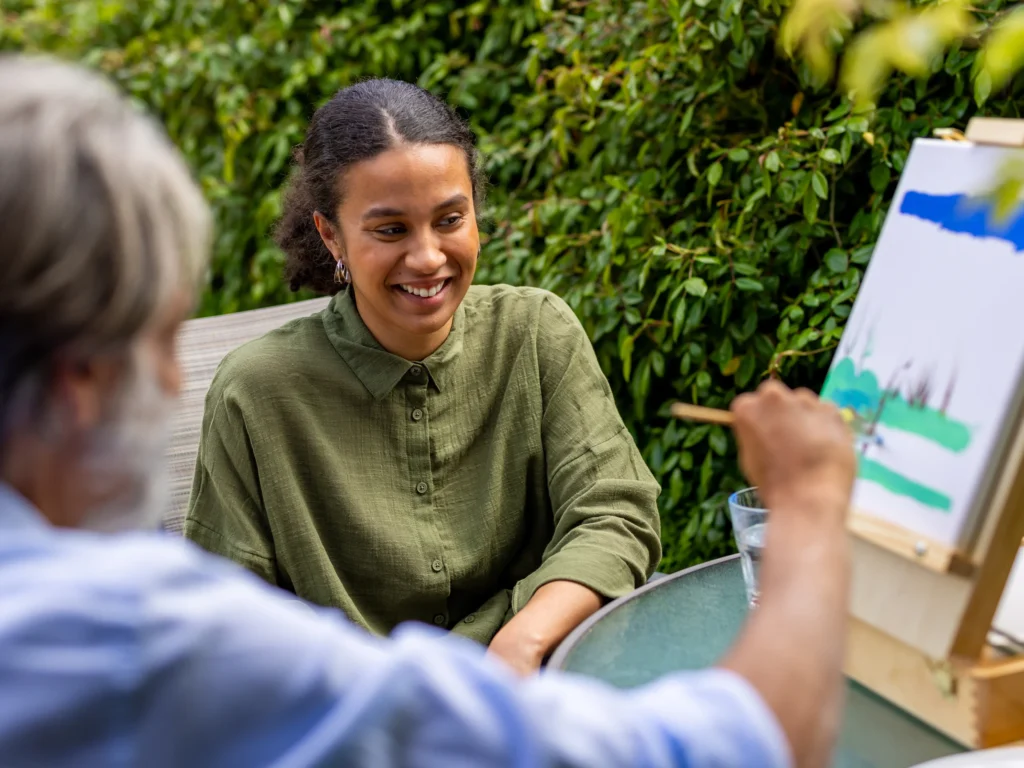 A young woman smiles whilst sitting at a glass table outdoors, watching another person paint on a canvas with a brush. Lush green foliage is visible in the background.