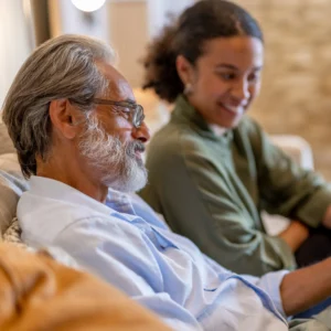 An older man with grey hair and glasses sits smiling on a sofa next to a young woman with curly hair, who is also smiling. They appear to be enjoying a pleasant conversation in a cosy indoor setting.