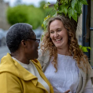 Two women smile and laugh together outdoors, standing close to each other near green leafy plants. One has curly hair and wears a white top; the other has short hair and wears glasses and a yellow jacket.