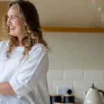 Two women, one older with short grey hair and glasses, and one younger with curly brown hair, smile and hold hands in a bright kitchen with white tiles and a vase of flowers on the worktop.