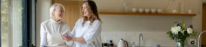 Two women, one older with short grey hair and glasses, and one younger with curly brown hair, smile and hold hands in a bright kitchen with white tiles and a vase of flowers on the worktop.