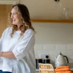 Two women, one older and one younger, smile and hold hands in a bright kitchen with fresh vegetables on the worktop and a vase of flowers nearby.