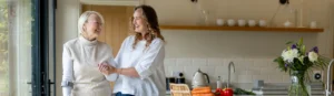 Two women, one older and one younger, smile and hold hands in a bright kitchen with fresh vegetables on the worktop and a vase of flowers nearby.