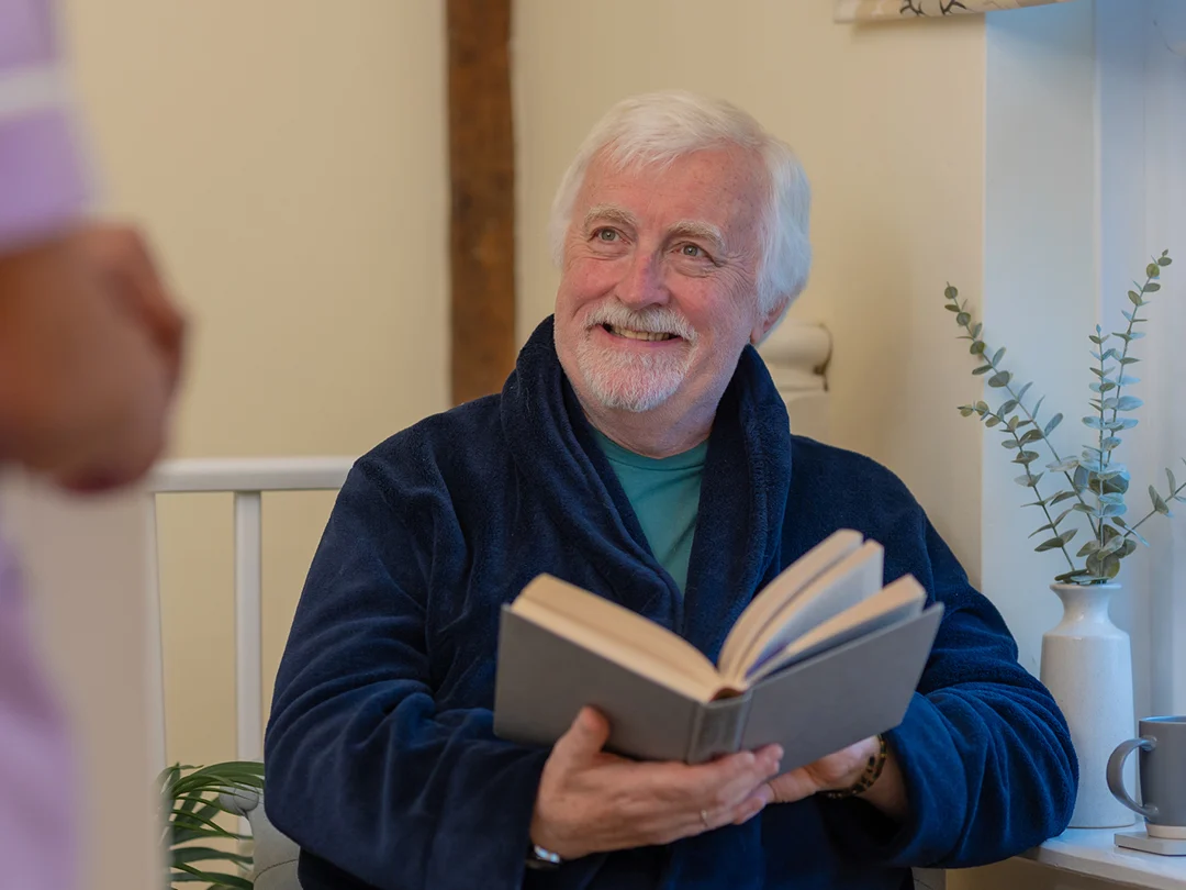 An older man with white hair and a beard, wearing a navy blue dressing gown, smiles whilst holding an open book. He sits indoors on a light-coloured chair with a plant and a vase in the background, looking at someone standing nearby.