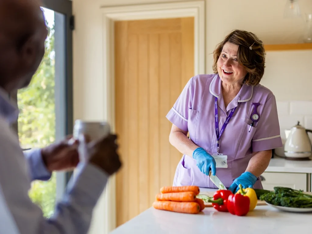 A female carer in a purple uniform and gloves smiles whilst preparing vegetables in a kitchen, including carrots, tomatoes, and a pepper, as an older adult watches from the foreground holding a mug.