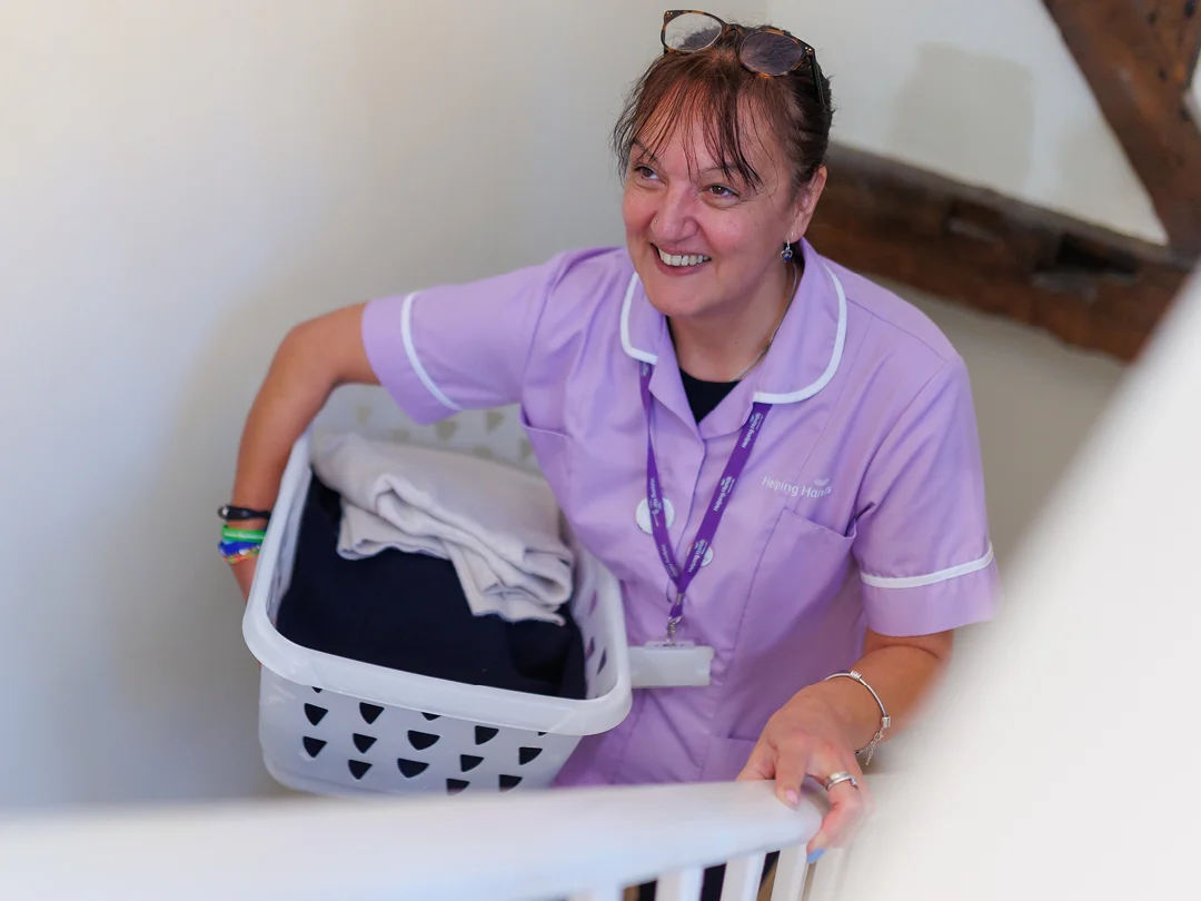 A woman in a purple uniform smiles while carrying a laundry basket with folded clothes up a staircase. She has glasses on her head, wears a lanyard, and appears to be a carer or healthcare worker.