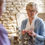 An older woman with short blonde hair and glasses smiles while knitting, sitting on a cushioned chair indoors. She wears a light blue cardigan and is engaged in conversation with another person.