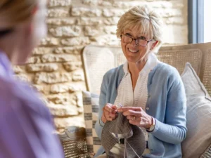 An older woman with short blonde hair and glasses smiles while knitting, sitting on a cushioned chair indoors. She wears a light blue cardigan and is engaged in conversation with another person.