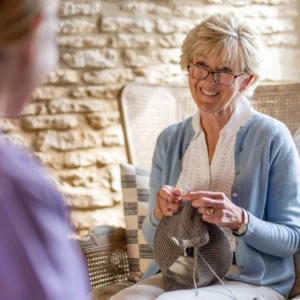 An older woman with short blonde hair and glasses smiles while knitting a grey garment, sitting on a wicker chair indoors and talking to another person whose back is facing the camera.