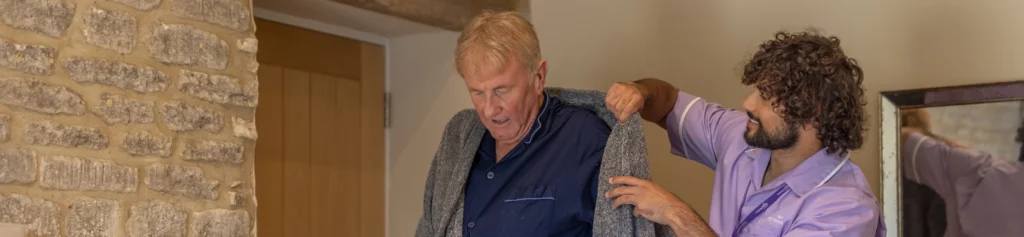 A carer helps an older man put on a grey cardigan in a cosy room with stone walls and a wooden door.