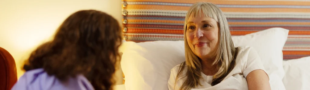 A woman with grey hair sits up in bed, smiling at another person with brown hair who is sitting nearby. The background shows a colourful striped headboard.