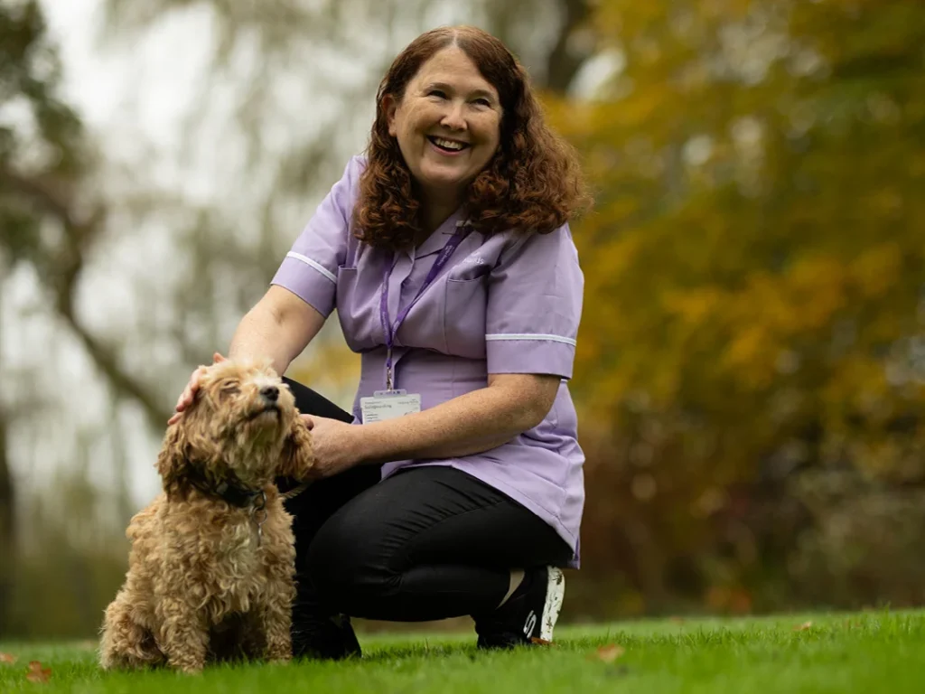 A smiling woman in a purple uniform kneels on grass, stroking a small, curly-haired dog. Trees with autumn-coloured leaves are blurred in the background.