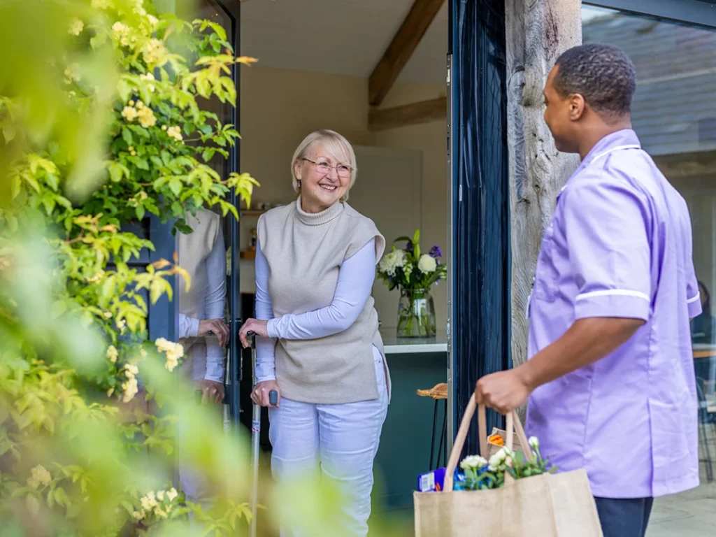 A smiling elderly woman opens her front door to a carer who stands outside, holding a shopping bag filled with food and flowers. The scene is bright and welcoming, with greenery visible in the foreground.