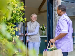 A smiling elderly woman opens her front door to a carer who stands outside, holding a shopping bag filled with food and flowers. The scene is bright and welcoming, with greenery visible in the foreground.