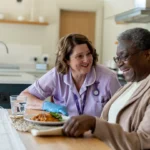 A nurse in a purple uniform smiles and talks with an elderly person at a kitchen table. The elderly person is smiling, and there is a plate of food and a glass of water in front of them.