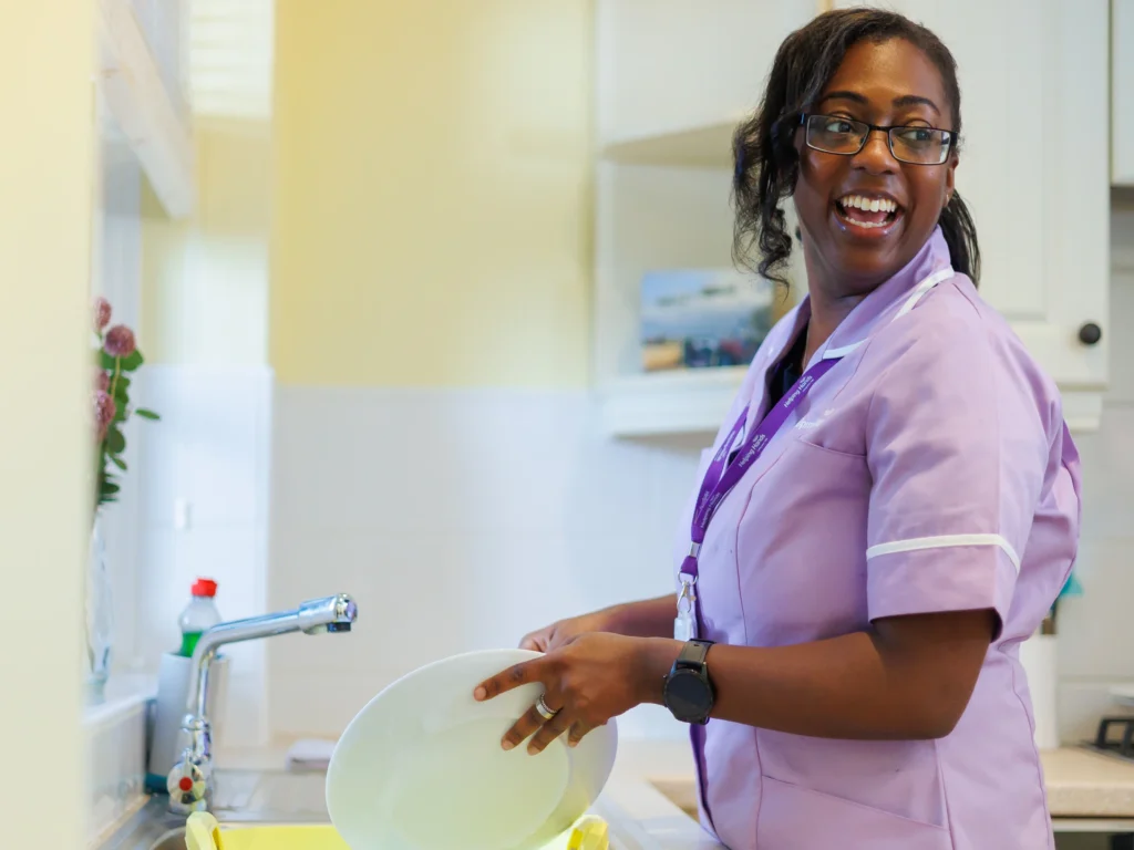 A woman in a light purple uniform smiles whilst washing up at a kitchen sink, holding a white plate. She wears glasses, a watch, and an ID lanyard. The kitchen is bright and tidy.