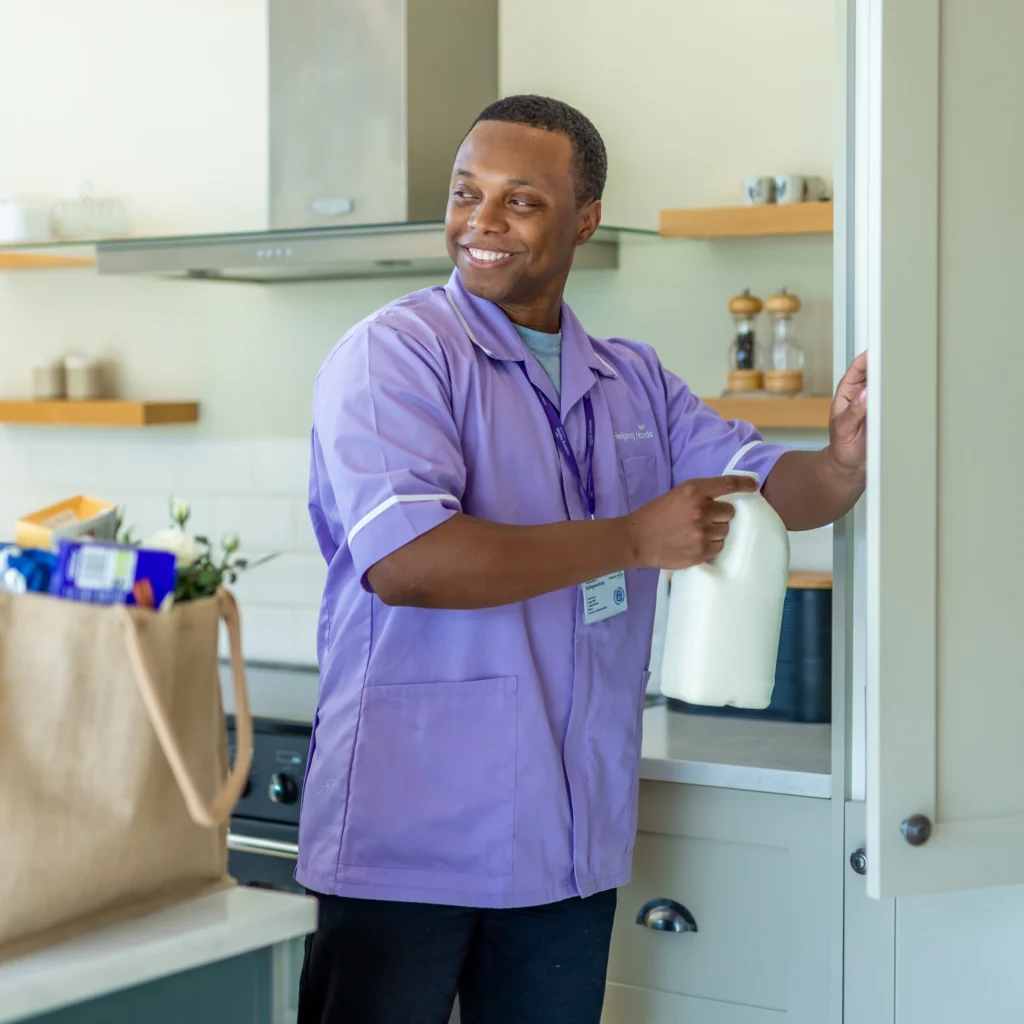 A man in a light purple uniform smiles while placing a bottle of milk in a kitchen cupboard. A reusable shopping bag filled with groceries sits on the worktop beside him.