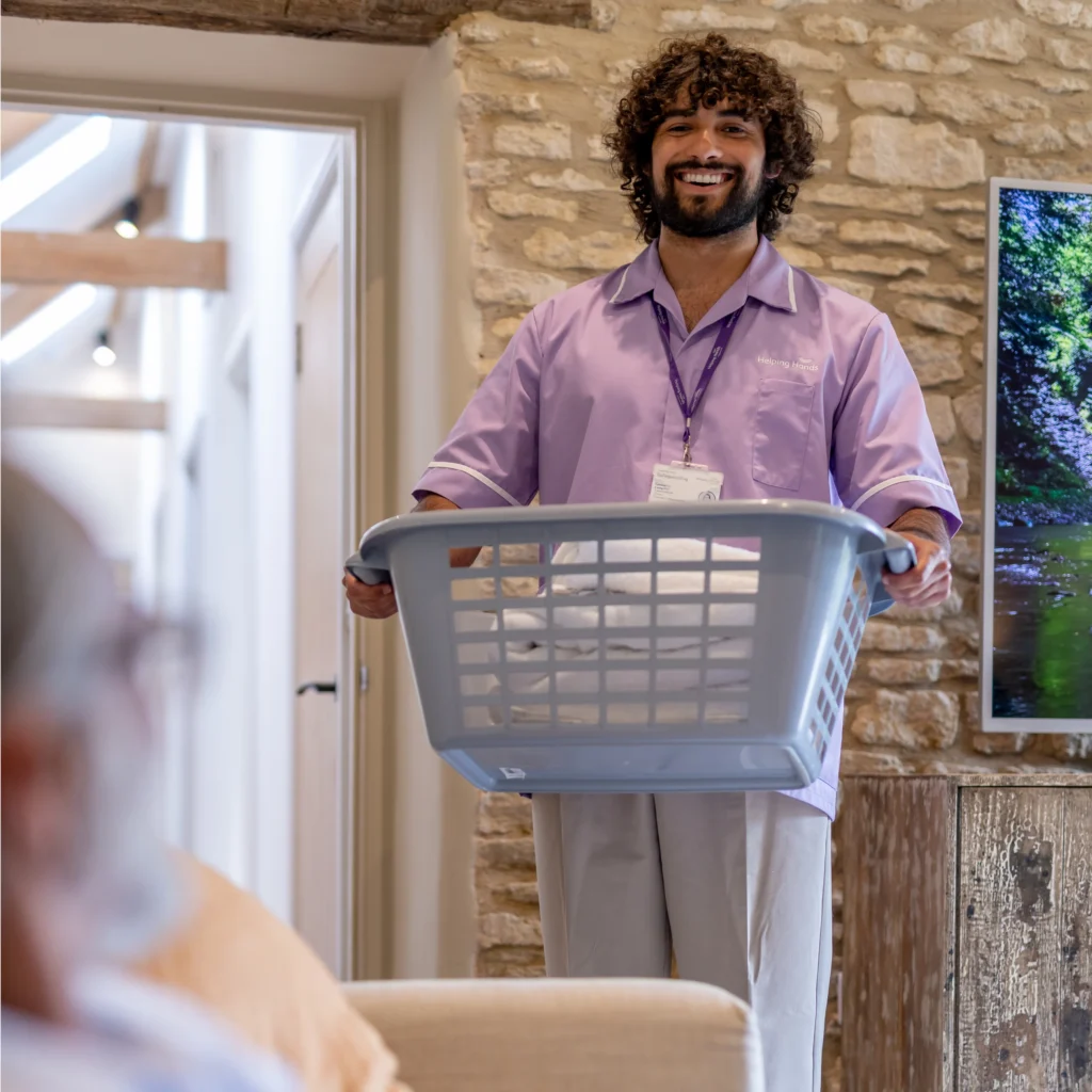 A smiling man wearing a purple uniform and lanyard holds a laundry basket with folded towels in a cosy, stone-walled room, whilst someone sits in the foreground.