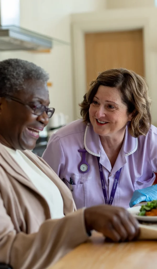 A smiling healthcare worker in a purple uniform sits beside an elderly woman, who is also smiling, as they share a moment together at a table with a meal.