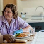 A nurse wearing gloves smiles and talks with an older adult at a kitchen table. The older adult is eating a meal, and there is a glass of water nearby. The atmosphere appears warm and friendly.